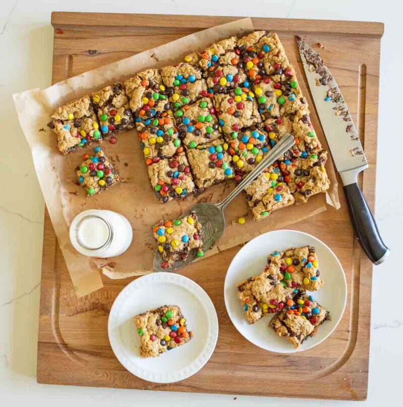 A wooden cutting board with cookie bars topped with colorful M&M candies, some cut into squares. A metal spatula, a knife, a glass of milk, and plates with more bars are also on the board.