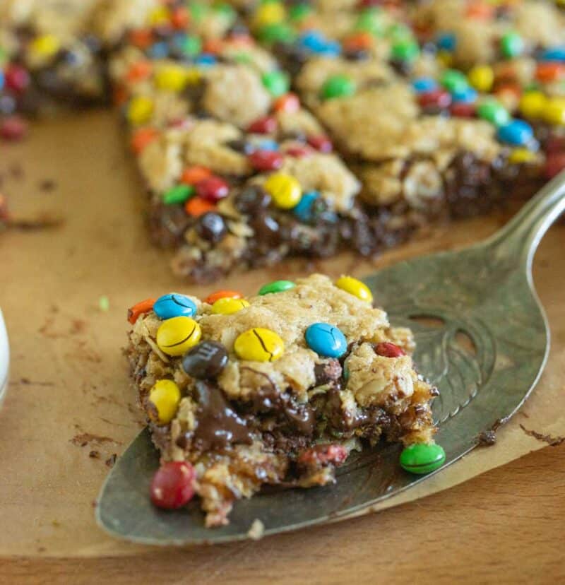 A close-up of a spatula holding an oatmeal cookie bar topped with colorful candy-coated chocolates and melted chocolate, with more bars in the background on parchment paper.