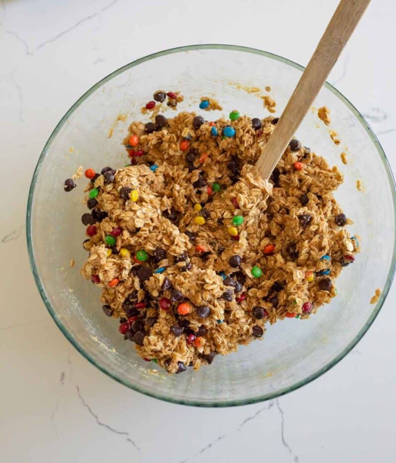 A glass bowl filled with oatmeal cookie dough mixed with chocolate chips and colorful candy-coated chocolates, being stirred with a wooden spoon on a white countertop.