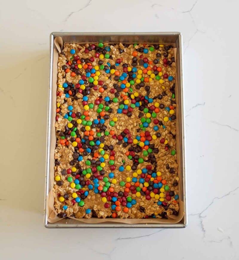 A rectangular baking pan filled with oatmeal bars topped with colorful mini chocolate candies and chocolate chips, on a white countertop.