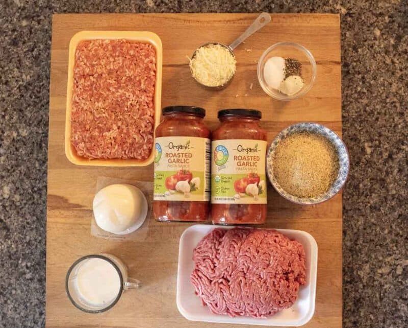 Ingredients for a recipe arranged on a wooden board, including ground meat, ground pork, two jars of roasted garlic pasta sauce, grated cheese, breadcrumbs, spices, an egg, and milk.