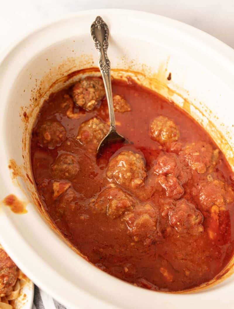 A white slow cooker filled with meatballs in red tomato sauce. A silver spoon is resting inside the cooker, partially submerged in the sauce and meatballs.