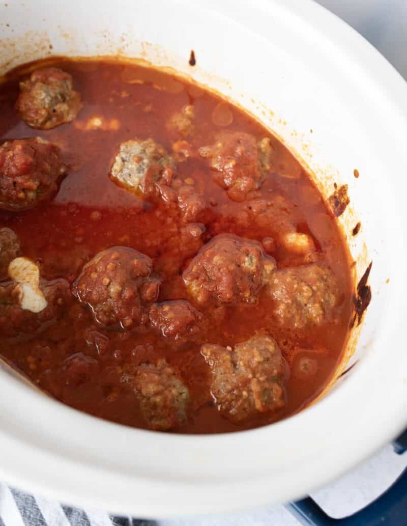 A white slow cooker filled with meatballs in rich red tomato sauce, some sauce splattered along the sides of the cooker.