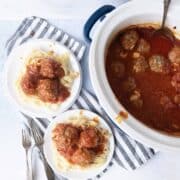 Overhead view of a slow cooker filled with meatballs in tomato sauce, next to two plates of pasta topped with meatballs and sauce, with forks placed beside the plates on a striped cloth.