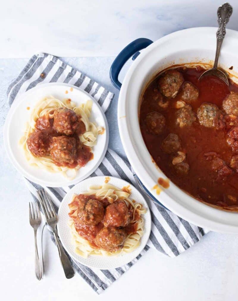 Overhead view of a slow cooker filled with meatballs in tomato sauce, next to two plates of pasta topped with meatballs and sauce, with forks placed beside the plates on a striped cloth.