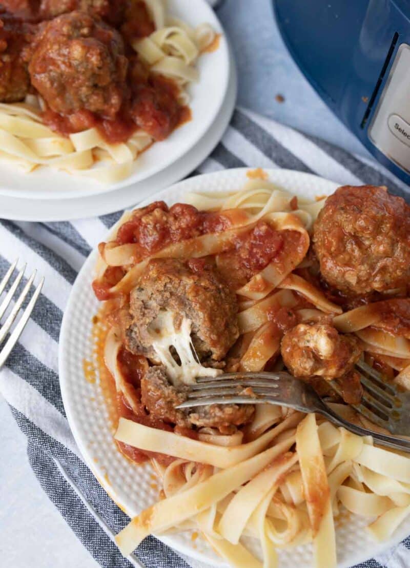 A close-up of a plate of fettuccine pasta topped with tomato sauce and meatballs. One meatball is cut open, revealing a melted cheese filling. A fork is resting on the plate beside the food.