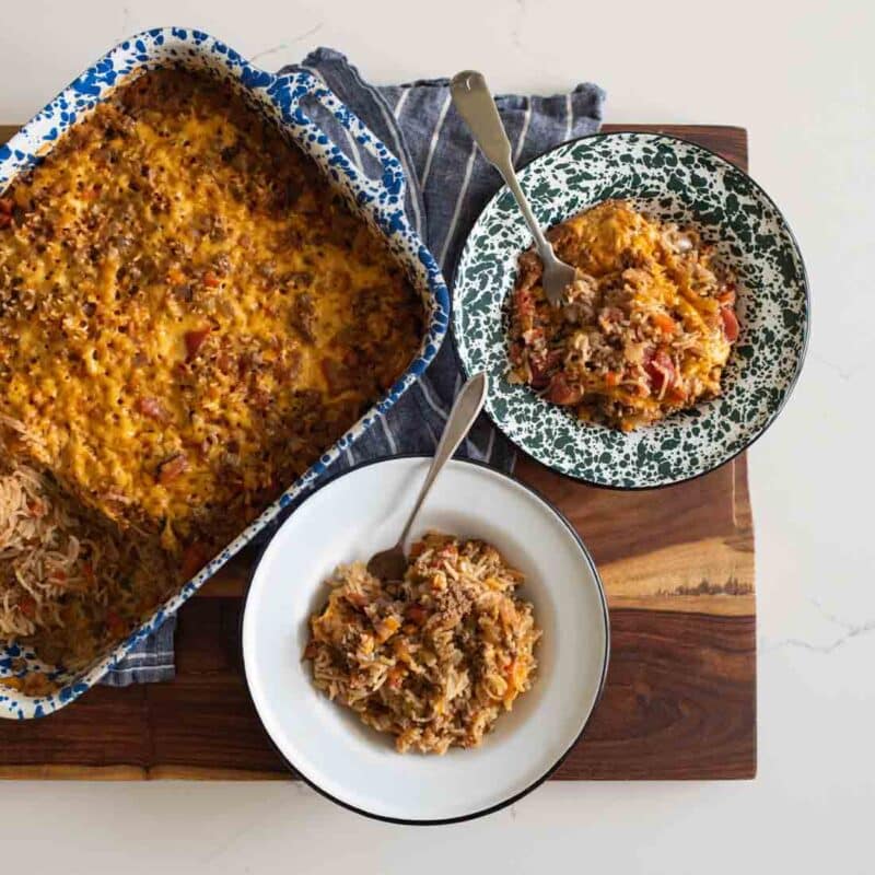 A casserole dish holds a cheesy baked rice and meat dish, with two servings scooped onto patterned plates beside it, all set on a wooden board with a striped cloth underneath.