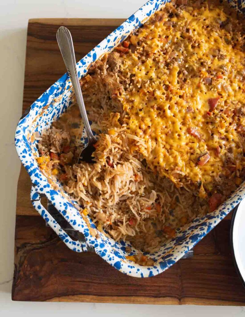A blue and white speckled baking dish filled with a cheesy rice casserole, partially served with a metal spoon resting inside. The casserole contains ground meat, shredded cheese, and vegetables.