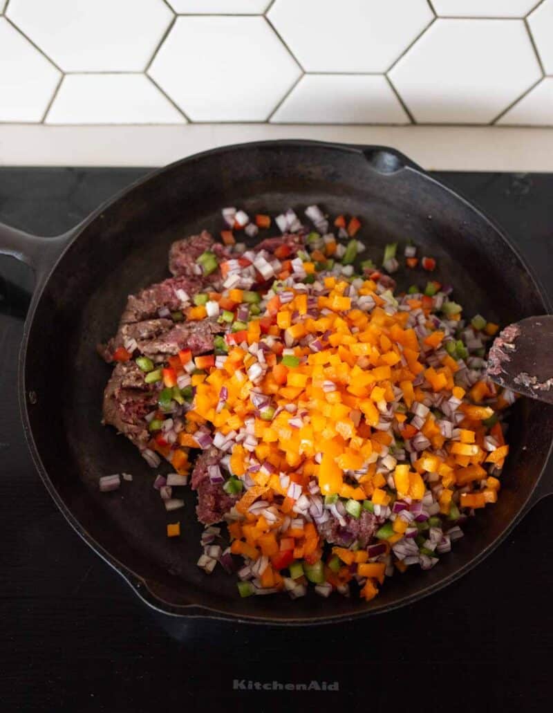Chopped red onion, orange bell pepper, green pepper, and raw ground beef in a black skillet on a stove, with a white hexagon tile backsplash in the background.