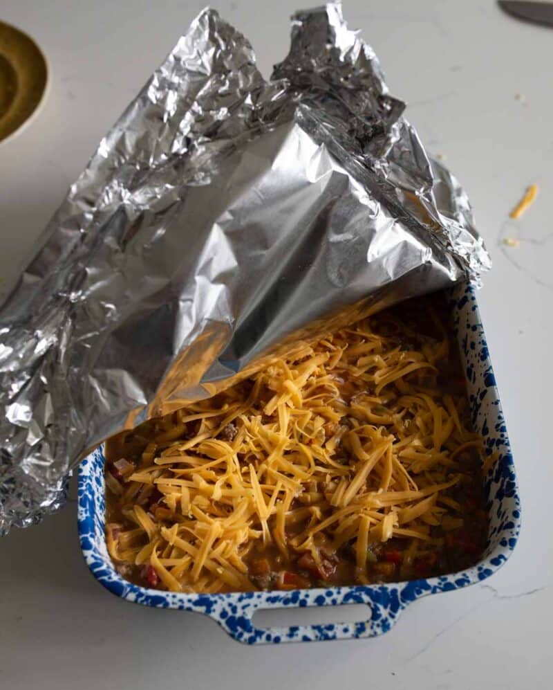 A blue and white speckled baking dish filled with shredded cheese on top of a casserole, partially covered by a sheet of aluminum foil.