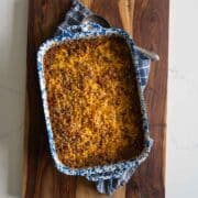 A baked casserole topped with melted cheese in a blue and white speckled dish, resting on a wooden cutting board with a blue plaid kitchen towel underneath.