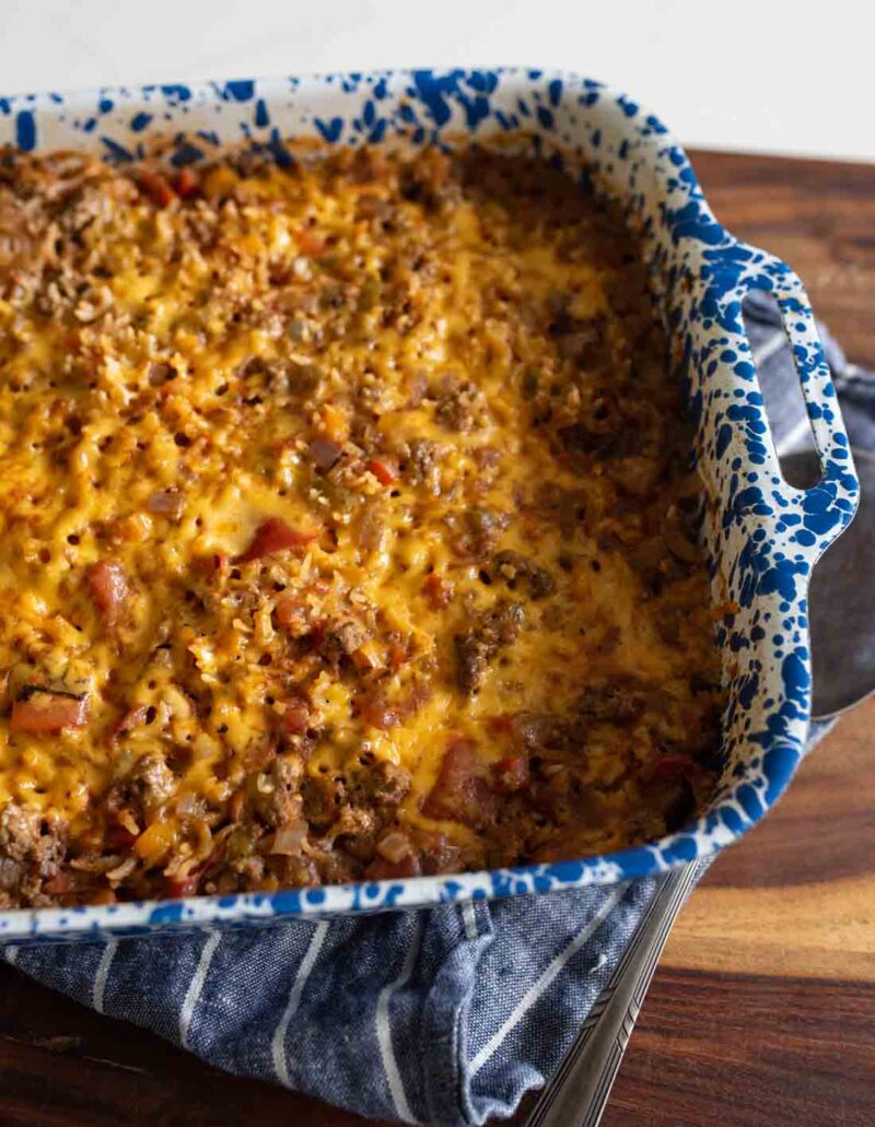 A blue and white speckled baking dish filled with a cheesy ground beef casserole sits on a wooden surface, partially resting on a striped towel.