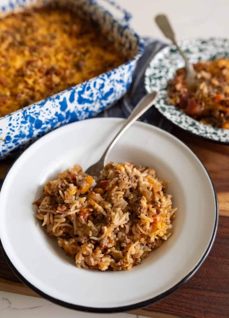 A serving of cheesy ground beef and rice casserole on a white plate with a fork, next to a baking dish filled with the same casserole and another plate of food in the background.